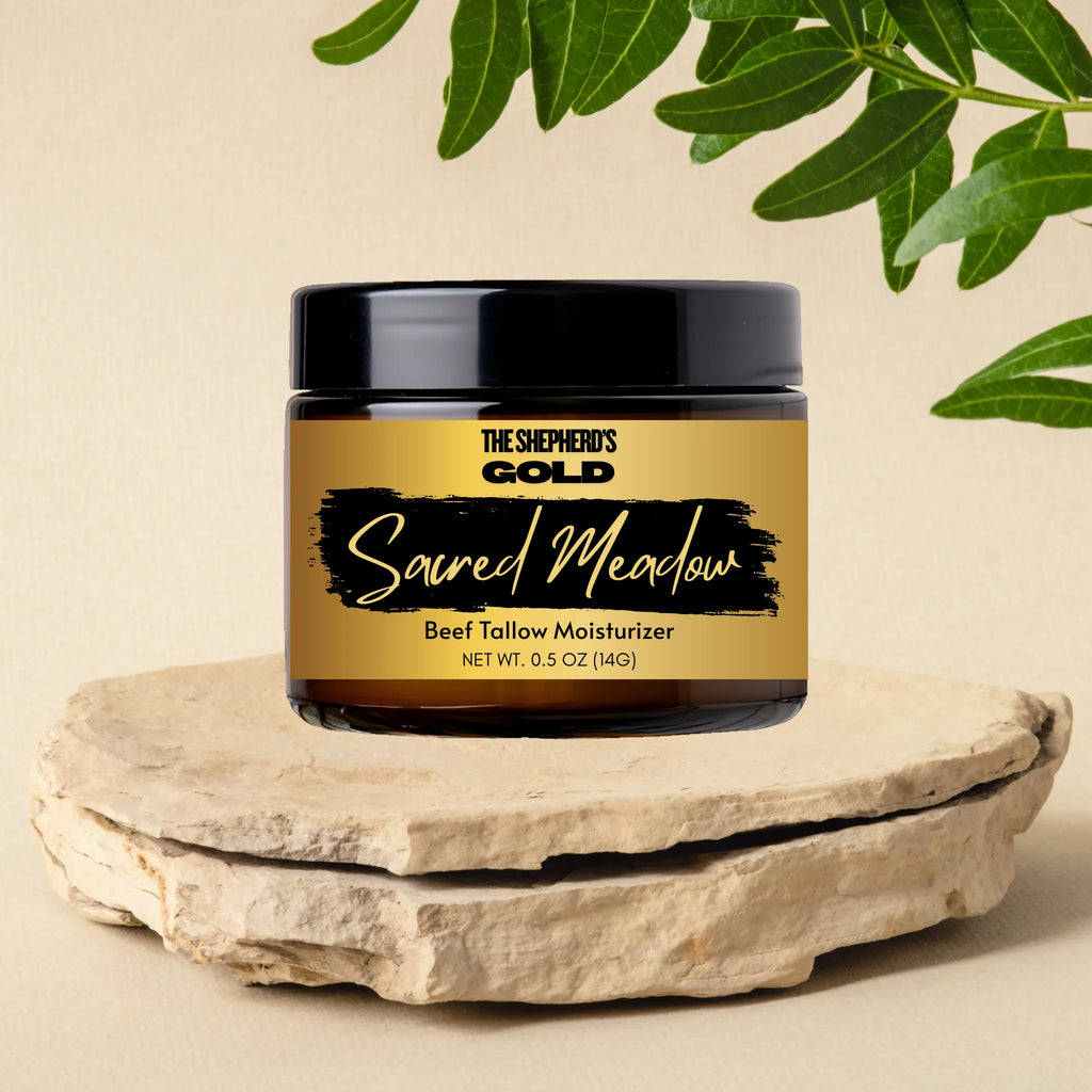 Jar of 'The Shepherd's Gold Sacred Meadow' beef tallow moisturizer on a stone surface with green leaves in the background.